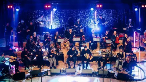 Kris Kesiak A large group of musicians play on stage with bright lights at Celtic Connections opening night.