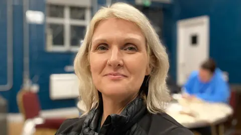 A woman with blonde shoulder length hair smiles toward the camera, she is wearing a black jacket. She is standing in a room with blue walls. In the blurred background, a person in a pale blue top sits writing at a desk.