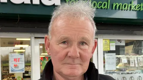 Shows head and shoulders of a man with grey hair standing in front of a supermarket