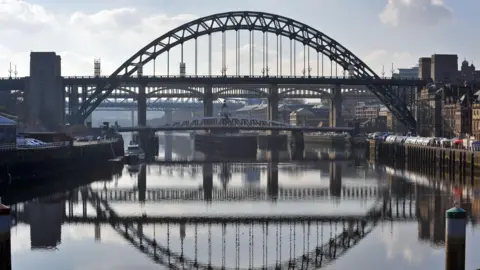 Picture of the Tyne Bridge, that stretches over the River Tyne in North East England, linking Newcastle upon Tyne and Gateshead. Pictured on a clear day with the bridge reflected in the river below.