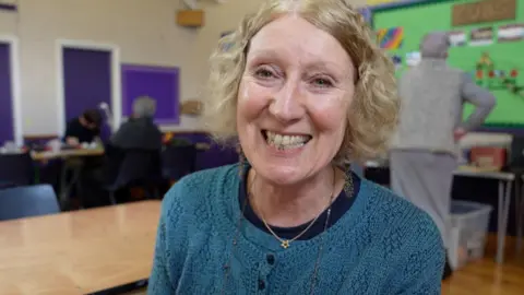 A woman with blonde hair and a blue cardigan and a small golden star necklace smiles as she sits in front of volunteers working in Pocklington Scout Hut.