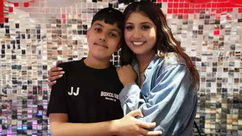 A woman wearing a denim jacket stands with her arm around a boy wearing a black t-shirt. The t-shirt has the letter 'JJ' and the BoxClever mentoring logo at the front.