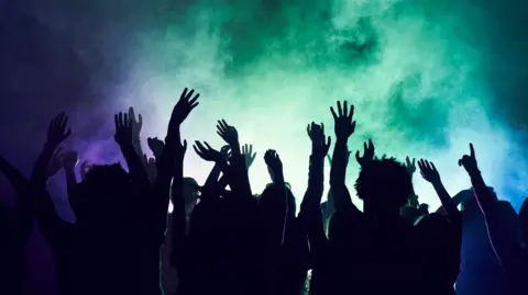 An image showing the silhouettes of people dancing in a nightclub. There is purple, green and blue coloured smoke in the air above.