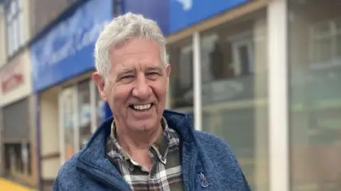 BBC/Claire Hamilton Nigel Nieto, who has short grey hair and is wearing a blue fleece above a check shirt, smiles while standing by a shopfront. 