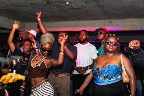 TEMILOLUWA JOHNSON / GETTY IMAGES Men and women dance inside a club. Out of shot is a DJ playing music on decks.