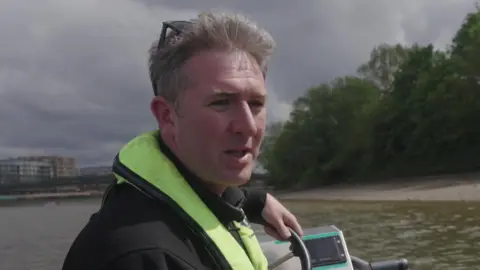 A man wearing a black jacket and a high-visibility yellow life vest sits in a small boat, holding the steering wheel with his left hand. The boat is on a river with a tree-lined bank in the background under a cloudy sky.