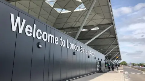 BBC The entrance to Stansted Airport, which has a sign welcoming visitors. It has a large white roof which is supported by metal beams. The entrance is next to a road with a zebra crossing.