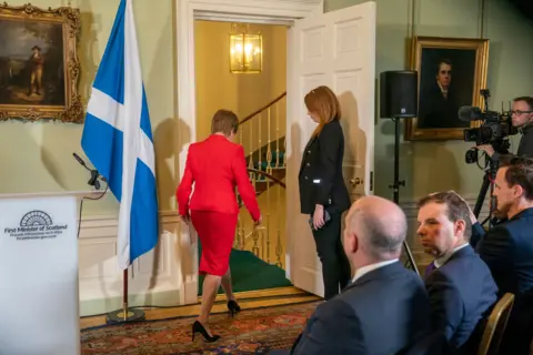 PA Media Nicola Sturgeon in a red suit walking out of a room full of journalists. Glenn Campbell and James Cook can been seen seated in the foreground. 