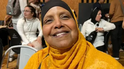A woman wearing a black and gold head scarf is smiling widely at the camera
