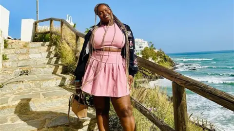 Handout Love Island fan Dami Amoo wears a pink two-piece set. She is standing on steps by coastline beside the ocean. 