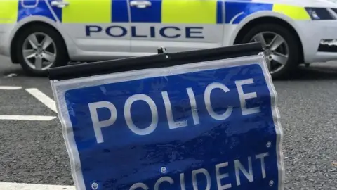 A blue police accident sign resting on the road in the foreground. In the background there is a parked police car with blue and yellow hi-vis decals.