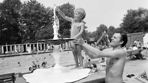 A black and white photo of a dad holding up his son so he can touch a water fountain at Malvern Park in Solihull, in 1976.
