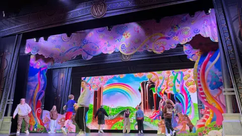 A number of children are on a stage in a theatre with their backs turned, standing in front of a colourful swirly background in the style of Charlie and the Chocolate Factory. There is a man wearing a cap, grey jumper and black jogging bottoms who is standing at the front of the stage. They appear to be rehearing for a production.