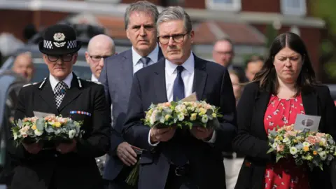 PA Media Merseyside Police Chief Constable Serena Kennedy, in full police uniform, Prime Minister Sir Keir Starmer, in a black suit, white shirt and black tie, and Merseyside Police and Crime Commissioner Emily Spurrell, in a red patterned dress and a black jacket, carry pink, peach and white floral wreaths