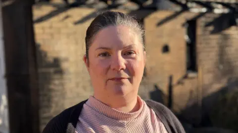 Kirsty Knight smiles at the camera. She has dark hair that is tied up behind her head and she is wearing a pink top with a black cardigan. The homes affected by the fire sit behind her.