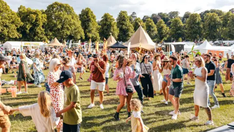 Shropshire Festivals A large crowd of people on a green grass field. They are dancing and holding drinks. In the background are various tall tents and flagpoles. There is a line of trees in the background.