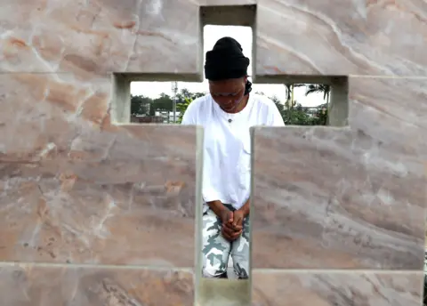 Legnan Koula / EPA A woman is seen throw the cross of gravestone as she bows her head and clasps her hands in Abidjan, Ivory Coast - Friday 1 November 2024