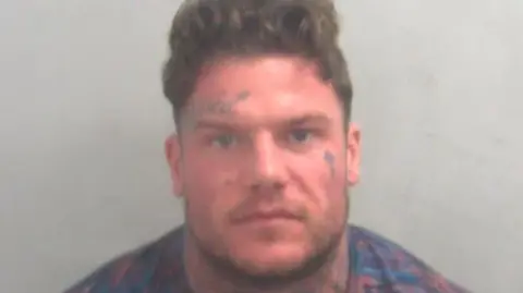Essex Police A custody image of Codey Nolan, who has brown hair and several face tattoos. He looks red in the face and annoyed.