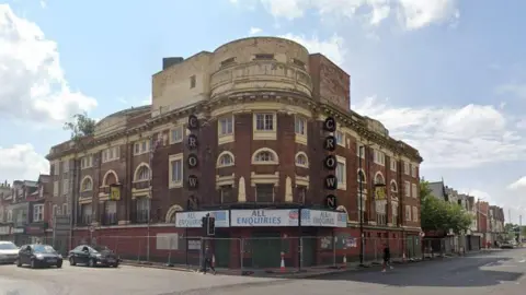 Google Maps image of the former Crown pub. It is a red-brick building with discoloured and peeling cream paintwork around the many windows. It stands on the corner of the street and is surrounded by metal fencing. Sale signs are above the main entrance.