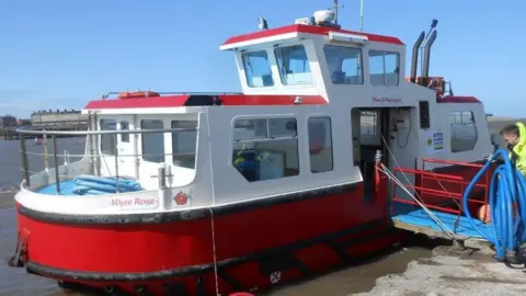 The Wyre Rose ferry, a red and white vessel with an enclosed seating area and a raised captain's cabin on top. A man is standing by the gangplank to the right in a high vis coat 
