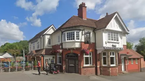 A large redbrick pub building on a sunny day. The building has two storeys and an outdoor beer garden area. A sign on the front of the building says "The Navigation Inn".