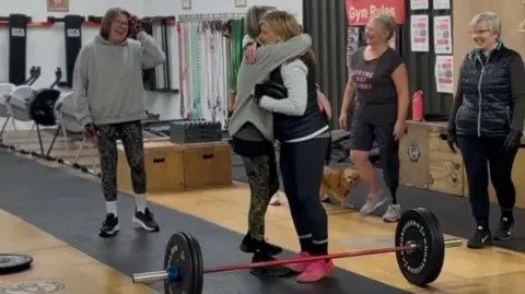Krish Stevens Women in a gym smiling and embracing. You can see a weightlifting bar on the floor and rowing machines in the background. 