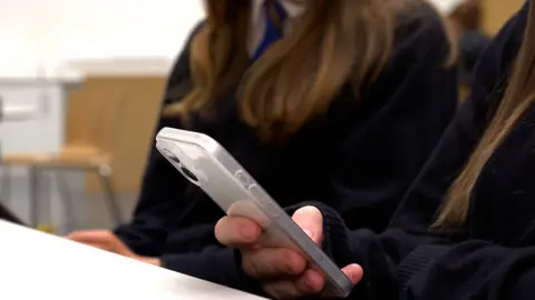 A close-up picture of a girl, whose face is off-camera, looking at a mobile phone while sat at a desk in school. Another girl, who is also unidentifiable, is sitting next to her. They are both wearing black jumpers.