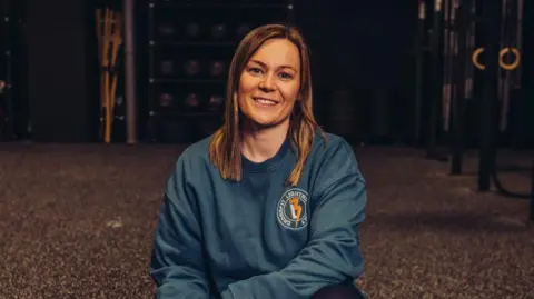 A woman with long fair hair and a blue jumper with a left chest logo is kneeling on the floor of a gym.