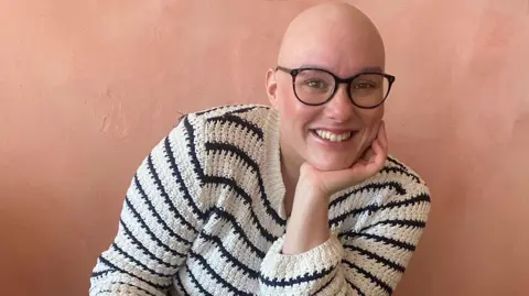 Laura Mathias Laura wears glasses and a cream jumper with black stripes. She is looking at the camera and smiling with her chin resting in her left hand.