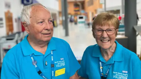 Shariqua Ahmed/BBC Ronald Nash has white hair and is wearing a blue polo shirt. He is standing next to Mary, who has short blonde hair and is wearing a matching polo shirt, and is smiling for the camera.
