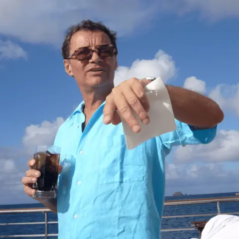 US Department of Justice A man in a short-sleeved, light blue shirt and sunglasses, holding a drink in one hand and a piece of paper in the other hand. He has short dark hair. A large expanse of water and blue sky with white clouds can be seen in the background. 