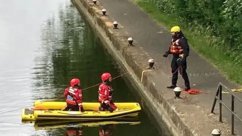 Two people wearing red gear in a yellow boat in a river, with one person stood wearing all black on the bank.