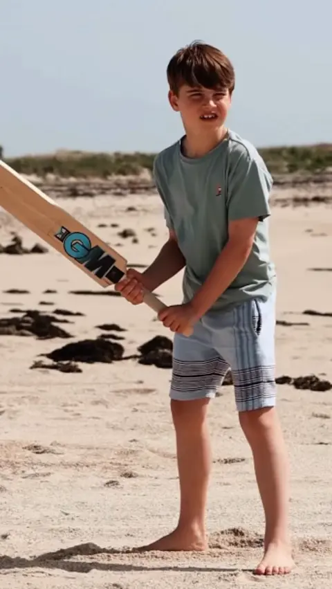 Matt Porteous Prince Louis squints while holding a cricket bat on a beach