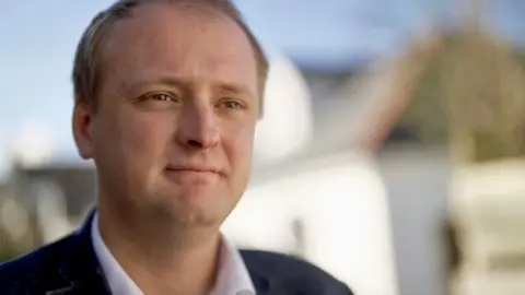 Stephen Fildes / BBC Ben Lake looks to the side of the camera. He has short, fair hair, wearing a dark suit and an open-necked white shirt. He is pictured outside with a white-walled building behind him. It is a sunny day. 