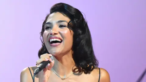 PA Media Olivia Dean, holding a microphone, on stage as she performs. She has long dark hair.