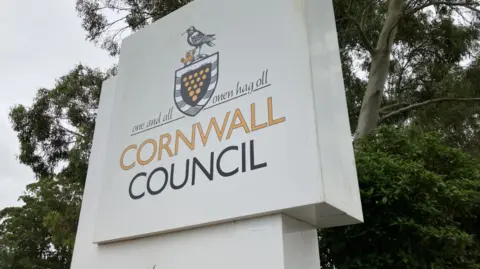 Close up of Cornwall Council sign. The white sign states Cornwall Council in yellow and black writing. A number of green trees are behind the sign.