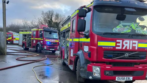 Three fire engines in a car park. Long hose reels lead off camera.