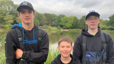 Kai, Harvey and Jack smile at the camera, all wearing black jumpers with Kai and Jack wearing caps. Bitterwell Lake is seen in the background. 