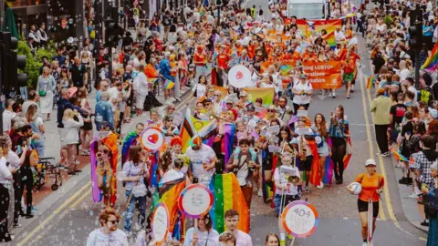 Leeds Pride parade with orange and rainbow banners, signs, floats, musicians, and bubbles watched on by people lining the streets