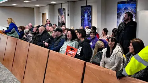 LDRS/Ian Duncan Dozens of people are sitting in the gallery of the council meeting, which consists of chairs lined behind a wooden panel. They make up three rows of chairs between them. Some are holding signs. One reads: "Together with refugees."