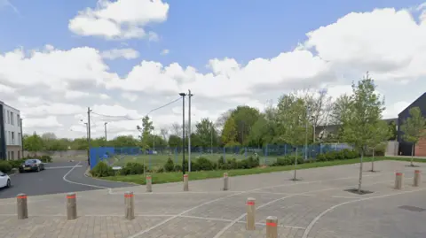 A concreate area with bollards leads to grass with a blue fence around it and parking to the left.
