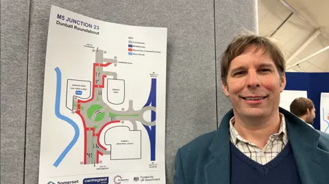 Councillor Richard Wilkins standing next to a poster about the planned works on the roundabout. He is wearing a blue sweater and a blue coat.