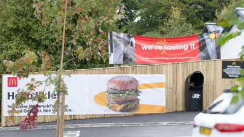 Rachel Laidler A red banner which reads 'we're not loving it' pokes up behind a wooden fence with a McDonald's advert on it. 
