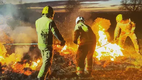 Three firefighters in full protection clothing and helmets are fighting a fire on a heath. It is just getting dark but there is still some flashes of sunlight in the sky 