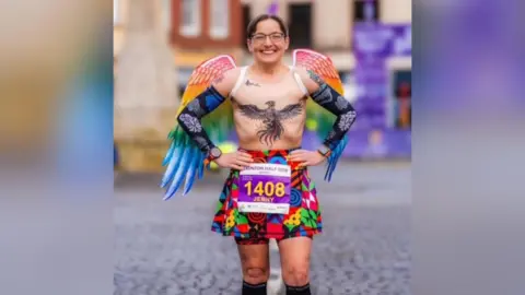 A woman stood with no top on, and a tattoo across her chest, while wearing multi-coloured wings and skirt, smiling at the camera.