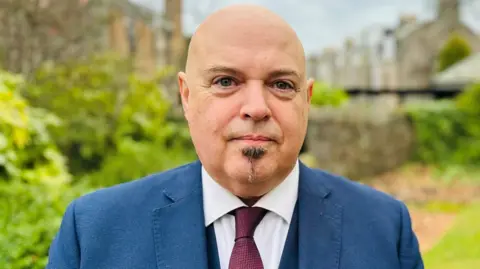 Councillor Richard Brooks, a man in a blue suit, white short and burgundy tie, looking at the camera.