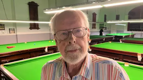 BBC/Phil Harrison Alastair Tod from the Royal Tunbridge Wells Civic Society stands in the snooker hall surrounded with tables and a snooker player in the background