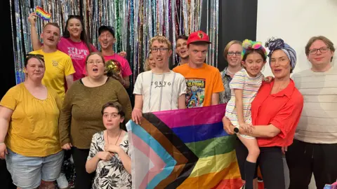 A group of 14 people, dressed in many colours, holding a pride flag in front of a glittery curtain 