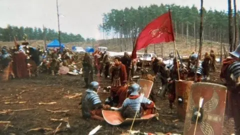 Scot Newing Bourne Woods, with a cast of extras dressed as roman soldiers, with flags, swords and shields. Cars and marquees are visible in the background, in front of a line of trees. 