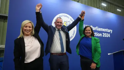 PA Media Cathal Mallaghan, centre, with Mary Lou McDonald holding up his left arm and Michelle O'Neill holding up his right arm